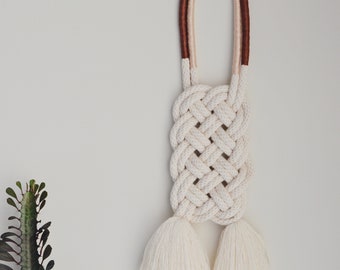 May include: A white macrame wall hanging with brown and pink accents. The hanging features a square knot pattern and two tassels at the bottom.