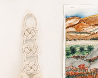 May include: A white macrame wall hanging with three knotted sections and two tassels at the bottom.