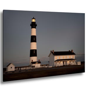 May include: A tall lighthouse with black and white stripes, topped with a lit lamp. A white house with a black roof and a small white building are next to the lighthouse. The sky is a dusky blue.