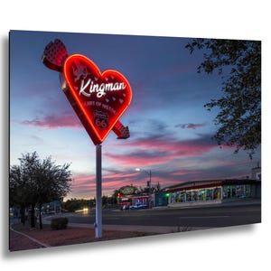May include: A neon sign in the shape of a heart pierced by an arrow, reading "Kingman Heart of History Route 66." The sign is red with white lettering, set against a dusk sky. A road and buildings are visible in the background.