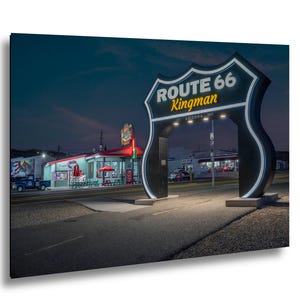 May include: A nighttime photograph of the Route 66 Kingman sign in Arizona. The sign is a large, black and white archway with the words "Route 66 Kingman" in white and yellow. A diner with red neon lights is in the background.