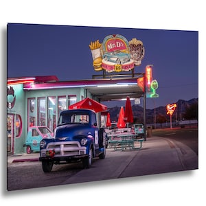 May include: A vintage blue pickup truck parked in front of Mr. D'z Route 66 Diner. The diner has a retro sign with neon lights and a red umbrella. A small pink car is parked nearby.