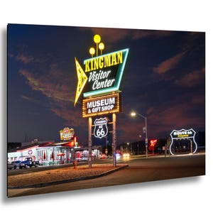 May include: A nighttime photograph of the Kingman Visitor Center sign, Museum & Gift Shop, and Route 66 sign in Kingman, Arizona. The signs are illuminated with neon lights against a dark blue sky. The image captures the essence of a classic American roadside attraction.