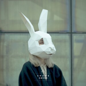 May include: A person wearing a white paper mache rabbit mask with the text "THE MASK SHAME COVER" on the bottom.