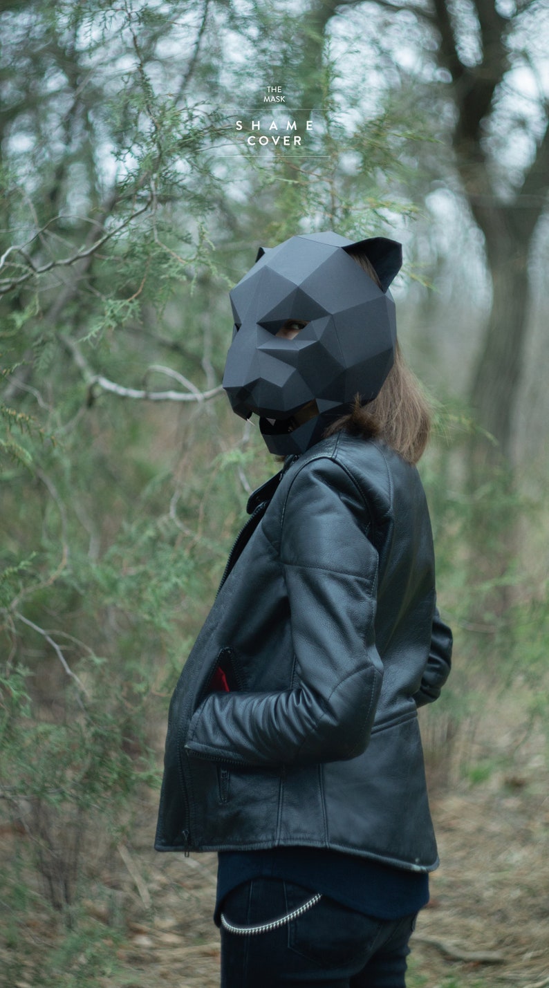 Op de afbeelding: Een persoon draagt een zwart leren jack en een zwart origami-dierenmasker. Het masker heeft een scherp, hoekig ontwerp en bedekt het gezicht van de persoon. De persoon staat in een bosrijke omgeving.