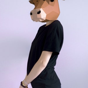 K&ouml;nnte beinhalten: Eine Person tr&auml;gt eine braune geometrische Papier-Kuhmaske mit wei&szlig;en H&ouml;rnern, ein schwarzes T-Shirt und eine schwarze Hose. Die Maske ist der Mittelpunkt, mit einem detaillierten, eckigen Design. Die Person steht vor einem schlichten Hintergrund.
