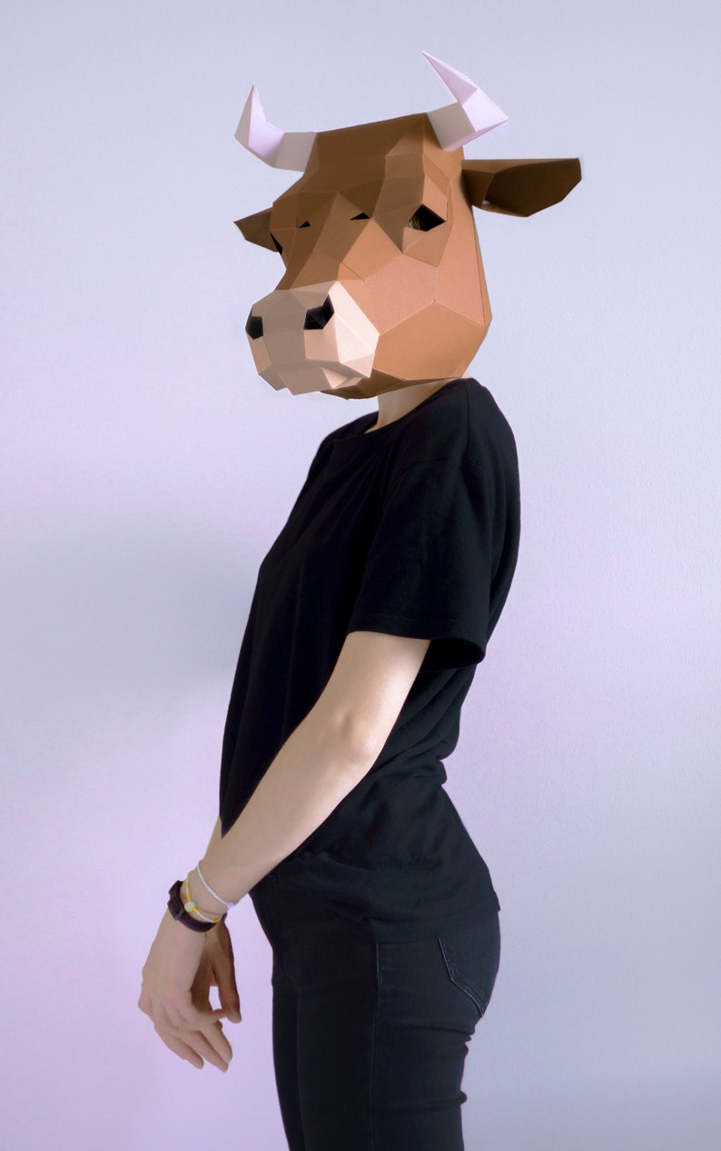 K&ouml;nnte beinhalten: Eine Person tr&auml;gt eine braune geometrische Papier-Kuhmaske mit wei&szlig;en H&ouml;rnern, ein schwarzes T-Shirt und eine schwarze Hose. Die Maske ist der Mittelpunkt, mit einem detaillierten, eckigen Design. Die Person steht vor einem schlichten Hintergrund.