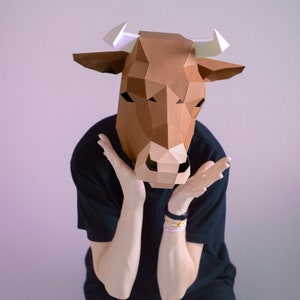 K&ouml;nnte beinhalten: Eine braune geometrische Papier-Kuhmaske mit wei&szlig;en H&ouml;rnern. Die Maske wird von einer Person getragen, die ein schwarzes T-Shirt und eine schwarze Hose tr&auml;gt. Die Person h&auml;lt ihre H&auml;nde vor sich.