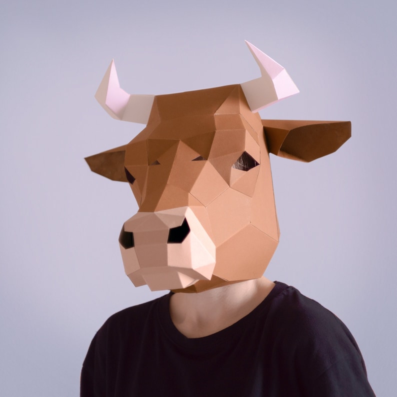 K&ouml;nnte beinhalten: Eine braune Papier-Kuhmaske mit wei&szlig;en H&ouml;rnern und einem geometrischen Design. Die Maske bedeckt das Gesicht und den Kopf, mit sichtbaren Augenl&ouml;chern. Die Person tr&auml;gt ein schwarzes Oberteil.