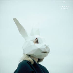 May include: A person wearing a white paper mask shaped like a rabbit's head. The mask has a geometric design and the person's eye is visible through a cut-out in the mask. The background is a light blue color. The text "THE MASK SHAME COVER" is printed in white at the top of the image.