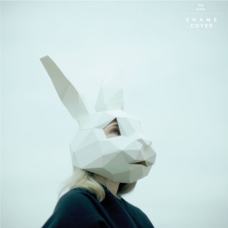 May include: A person wearing a white paper mask shaped like a rabbit's head. The mask has a geometric design and the person's eye is visible through a cut-out in the mask. The background is a light blue color. The text "THE MASK SHAME COVER" is printed in white at the top of the image.