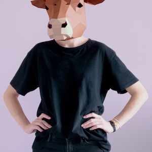 K&ouml;nnte beinhalten: Eine Person tr&auml;gt eine geometrische Kuhmaske in Braun und Wei&szlig;, ein schwarzes T-Shirt und eine schwarze Jeans. Die Person steht mit den H&auml;nden auf den H&uuml;ften. Der Hintergrund ist einfarbig hellviolett.