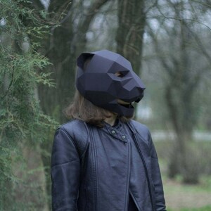Op de afbeelding: Een persoon draagt ​​een zwart origami-achtig dierenmasker in een bosrijke omgeving. Het masker is gevormd als een beer of een panter en heeft een geometrisch ontwerp.