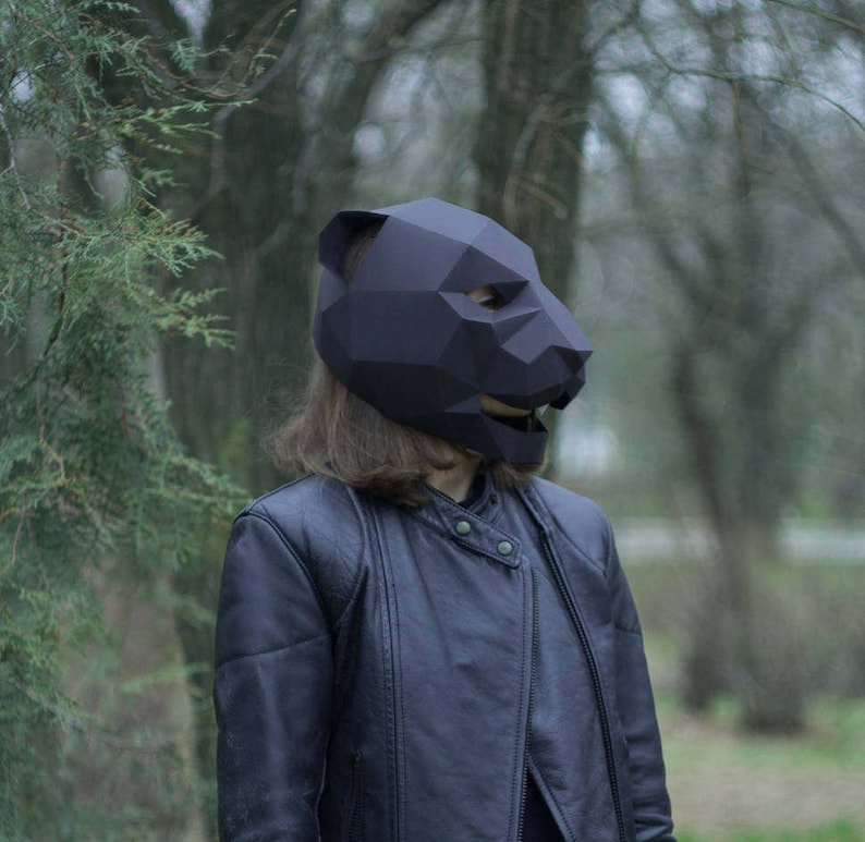 Op de afbeelding: Een persoon draagt ​​een zwart origami-achtig dierenmasker in een bosrijke omgeving. Het masker is gevormd als een beer of een panter en heeft een geometrisch ontwerp.