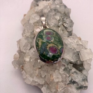 May include: A silver pendant featuring a green and pink oval gemstone with a silver bail. The pendant is resting on a bed of white crystals.