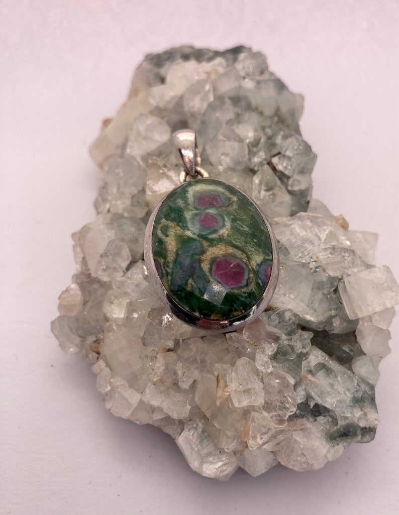 Puede incluir: Un colgante de plata con una piedra ovalada verde y rosa con una anilla de plata. El colgante est&aacute; apoyado en un lecho de cristales blancos.