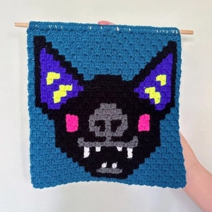 May include: A crocheted wall hanging featuring a black bat with purple and yellow ears and pink cheeks. The bat has a toothy grin and is set against a blue background.