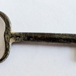 May include: A vintage-style key with a circular handle and a rectangular bit. The key is made of a dark, aged metal, possibly bronze or iron, with visible signs of wear and oxidation. The key's design suggests it may be antique or a replica.