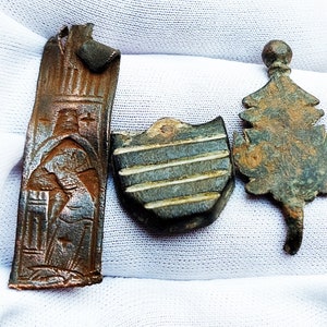 May include: Three antique bronze metal pieces: a rectangular plaque with a raised design of a person, a shield-shaped piece with a striped pattern, and a leaf-shaped piece with a textured surface.