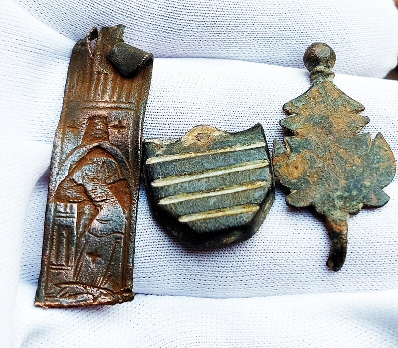 May include: Three antique bronze metal pieces: a rectangular plaque with a raised design of a person, a shield-shaped piece with a striped pattern, and a leaf-shaped piece with a textured surface.