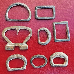 Może przedstawiać: Dziewięć antycznych srebrnych klamer metalowych o różnych kształtach i rozmiarach. Klamry są ułożone w losowy wzór na czerwonym tle.