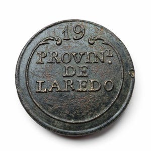 Op de afbeelding: Een ronde, antieke bronzen munt met de tekst "19 PROVIN DE LAREDO" in reliëf. De munt heeft een donkere, verouderde uitstraling met tekenen van slijtage, wat wijst op de leeftijd en geschiedenis. De tekst is in hoofdletters.