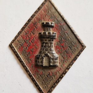 Puede incluir: Una insignia de metal en forma de diamante con fondo rojo y negro. La insignia presenta una imagen en relieve tridimensional de una torre de piedra con un techo puntiagudo.