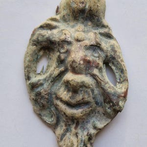 May include: A green and brown metal mask with a carved face and two holes on the sides. The mask is in the shape of a face with a wide mouth and a long nose.