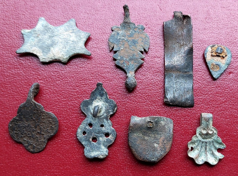 May include: A collection of eight antique metal pendants in various shapes and designs. The pendants are made of different metals, including silver, copper, and bronze. Some of the pendants have intricate details, while others are more simple. The pendants are all in good condition, despite their age.