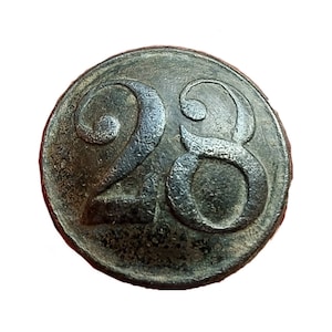 Op de afbeelding: Een ronde metalen knoop met het nummer "28" erop gegraveerd. De knoop is donkergrijs van kleur en heeft een licht verweerde uitstraling.