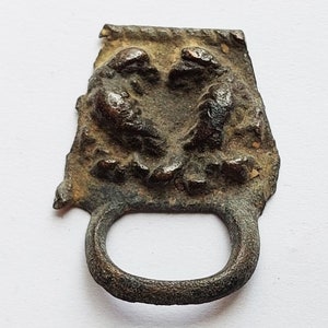 Puede incluir: Un artefacto metálico desgastado, de color bronce, con una parte superior rectangular y un lazo circular. La parte superior presenta un relieve detallado de dos figuras. La pieza tiene una apariencia envejecida, lo que sugiere importancia histórica.