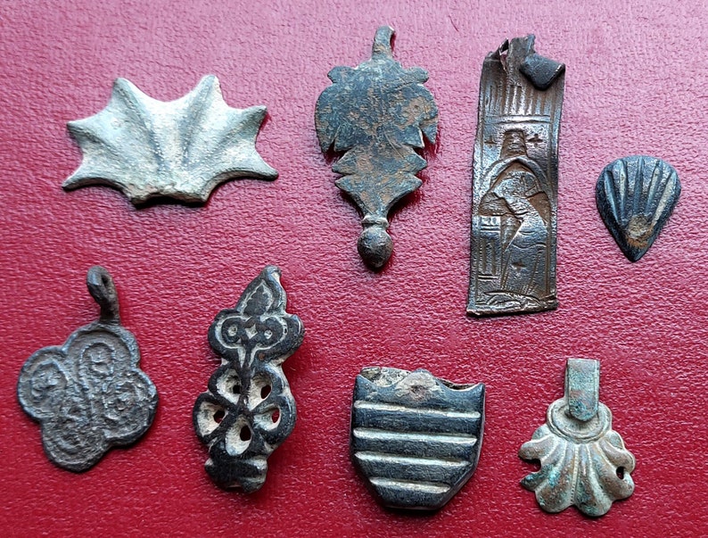 May include: A collection of nine antique metal pendants with various designs, including a star, a leaf, a religious figure, a shield, and a flower. The pendants are made of silver, bronze, and copper.