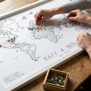 May include: A framed world map with a white border, featuring black outlines of continents and countries. The map is personalized with the names "KATY & BEN" and "EST 2019". Colorful push pins mark travel locations. A small wooden box holds additional pins.