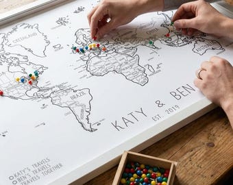 May include: A framed world map with a white border, featuring black outlines of continents and countries. The map is personalized with the names "KATY & BEN" and "EST 2019". Colorful push pins mark travel locations. A small wooden box holds additional pins.