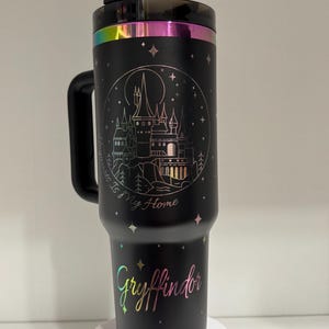 Puede incluir: Taza de viaje de acero inoxidable negro con asa. La taza tiene un acabado hologr&aacute;fico y presenta un gr&aacute;fico de un castillo con el texto "Home is where my heart is" y "Gryffindor".