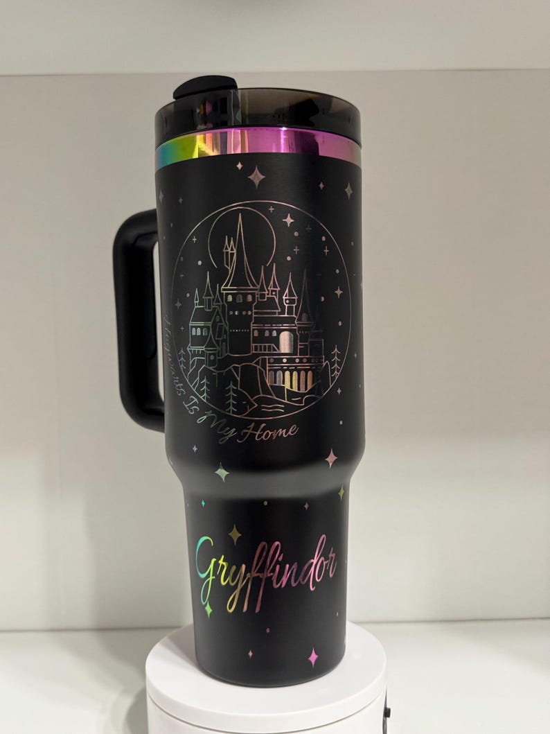 Puede incluir: Taza de viaje de acero inoxidable negro con asa. La taza tiene un acabado hologr&aacute;fico y presenta un gr&aacute;fico de un castillo con el texto "Home is where my heart is" y "Gryffindor".