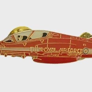 May include: A red and gold Royal Air Force jet pin. The pin features a detailed design of a jet aircraft with the words "ROYAL AIR FORCE" on the side. The jet has a gold canopy and accents.