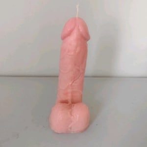 May include: A light pink wax candle moulded into the shape of a phallus. The candle features a smooth, matte texture with a single white cotton wick protruding from the top. The base consists of two rounded, symmetrical sections of wax.