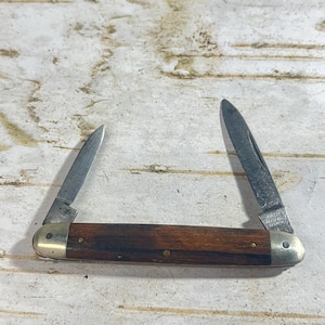 May include: A vintage pocket knife with two blades extended. The handle is made of dark brown wood with brass accents. The blades are silver and show signs of wear. The knife is open, displaying its functionality.