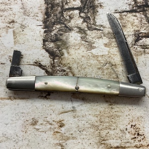 May include: A vintage pocket knife with a mother-of-pearl handle and two open blades. The blades are dark with signs of wear, and the handle has a pearlescent sheen. The knife is set against a textured background.