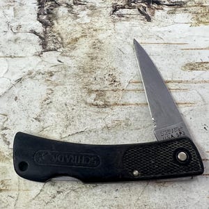 May include: A black folding knife with a silver blade. The handle is textured and features the brand name "SCHRADE" and the text "USA SP3". The knife is designed for outdoor use and everyday carry.
