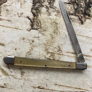 May include: A vintage pocket knife with a stainless steel blade and a light-colored handle. The blade is open, revealing the sharp edge. The handle has a brass-colored section. The knife is marked "Jabre Stainless Japan".