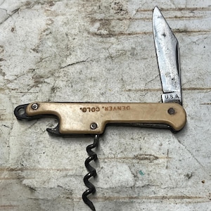 May include: A vintage multi-tool with a cream-colored handle, featuring a corkscrew, bottle opener, and a partially open blade. The blade is marked "U.S.A." and the handle reads "DENVER, COLO."