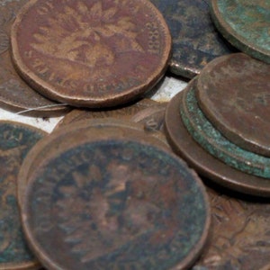 Pu&ograve; includere: Una collezione di antiche monete Indian Head, con un disegno dettagliato di un nativo americano con un copricapo di piume. Le monete sono di tonalit&agrave; marroni e verdi, con segni di usura visibili.