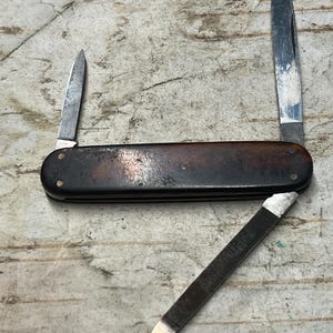 May include: A vintage pocket knife with a dark brown handle and three open blades. The blades are silver and show signs of wear. The knife is lying on a textured, light gray surface.