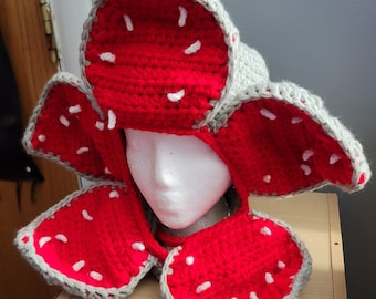 Puede incluir: Un gorro de crochet con forma de flor roja y blanca con detalles grises. El gorro está diseñado para parecerse al monstruo de la serie Stranger Things.