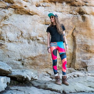 Op de afbeelding: Een vrouw in een zwart T-shirt, een blauwe en groene geprinte pet en kleurrijke leggings staat voor een rotsachtige klif.