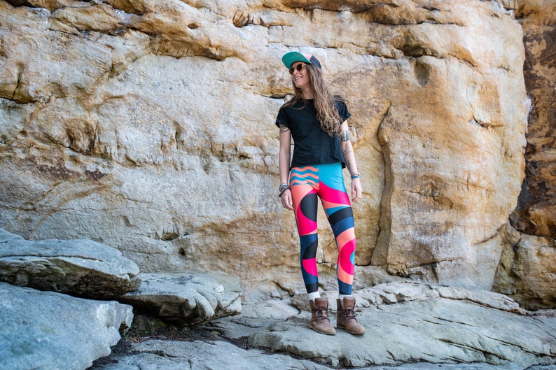 Op de afbeelding: Een vrouw in een zwart T-shirt, een blauwe en groene geprinte pet en kleurrijke leggings staat voor een rotsachtige klif.