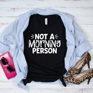Pu&ograve; includere: Maglietta nera con testo bianco che recita "Not a Morning Person" su uno sfondo di legno bianco. La maglietta &egrave; abbinata a una camicia a righe blu e bianche e scarpe leopardate.