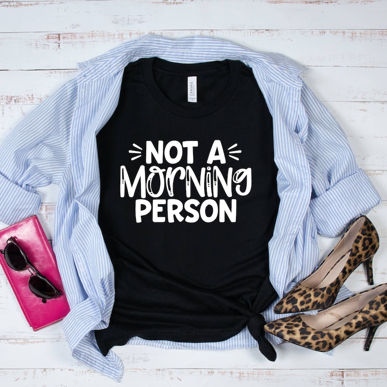 Pu&ograve; includere: Maglietta nera con testo bianco che recita "Not a Morning Person" su uno sfondo di legno bianco. La maglietta &egrave; abbinata a una camicia a righe blu e bianche e scarpe leopardate.